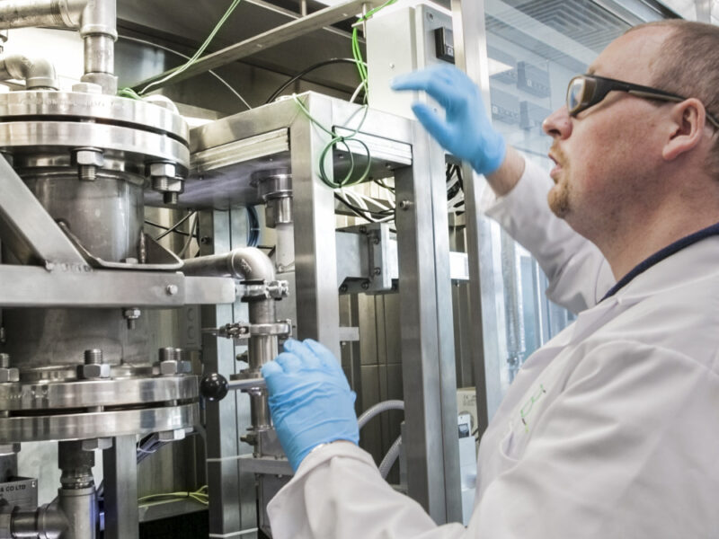 Scientist in lab coat and goggles assessing large scale processing equipment